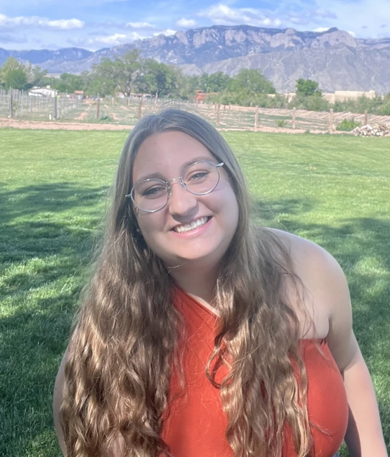Person with long blonde hair and glasses smiling on field with mountains in the background