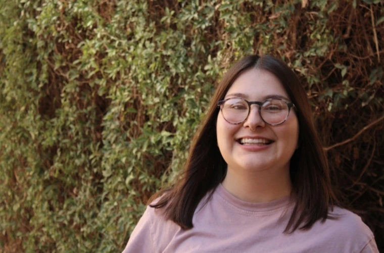 Person with glasses and shoulder length hair smiling in front of green plants