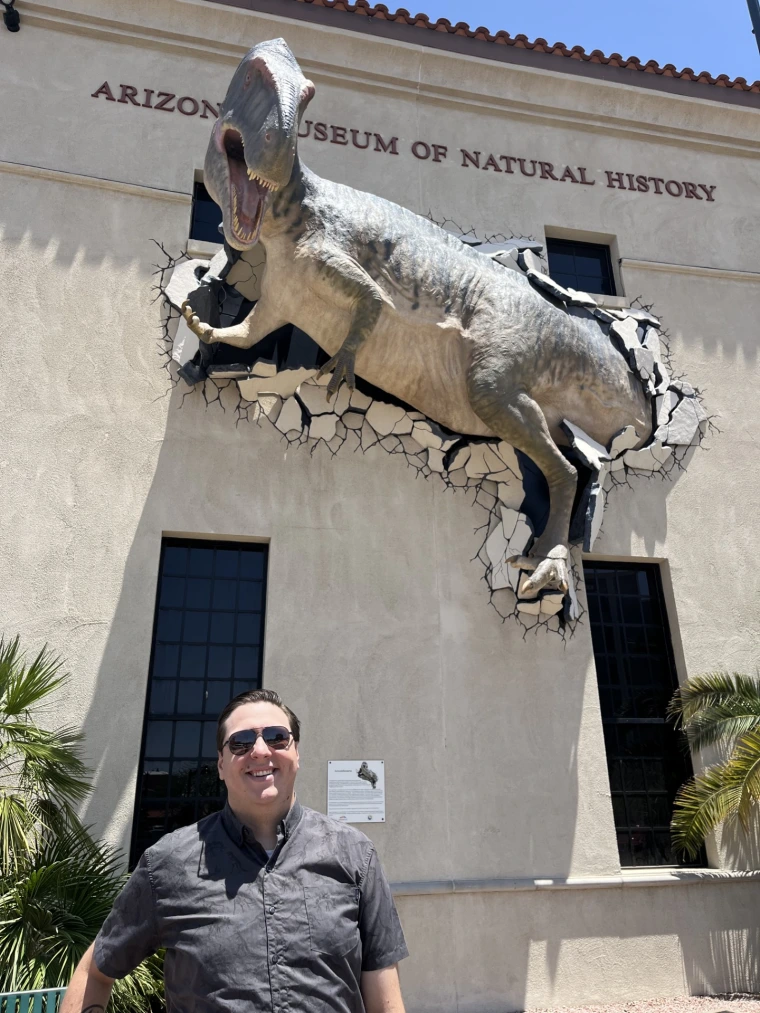 Person in black shirt and sunglasses smiling under dinosaur statue at the Arizona Museum of Natural History