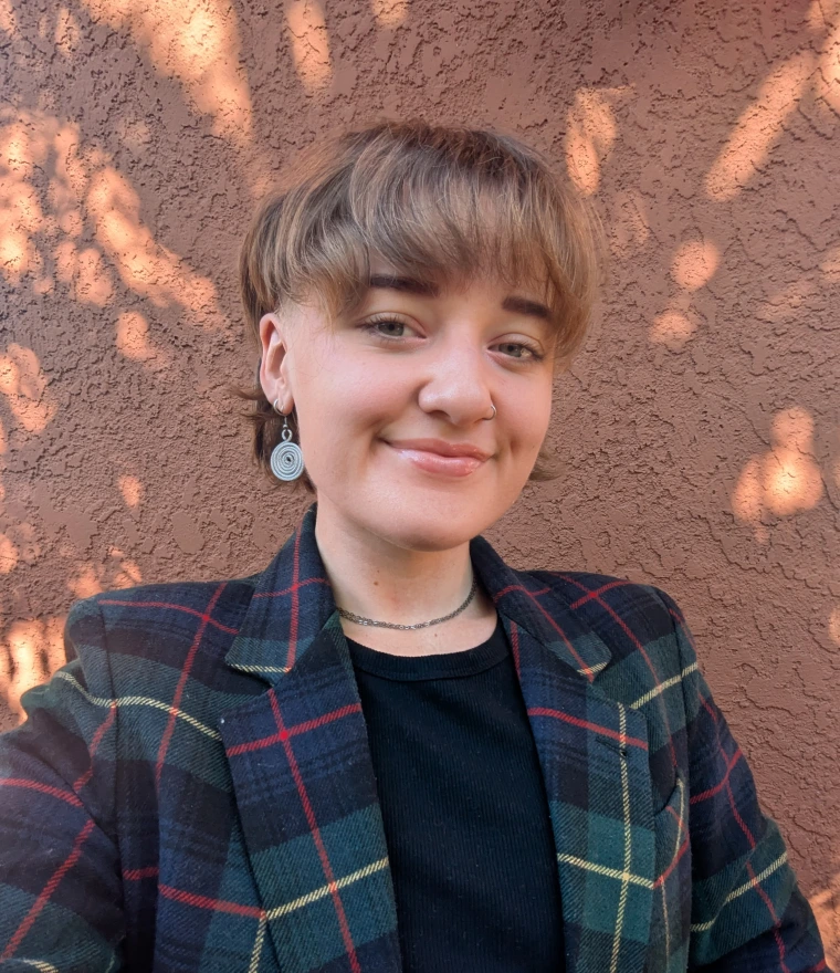 Selfie of person with short blonde hair in plaid blazer smiling at camera in front of terracotta stucco wall